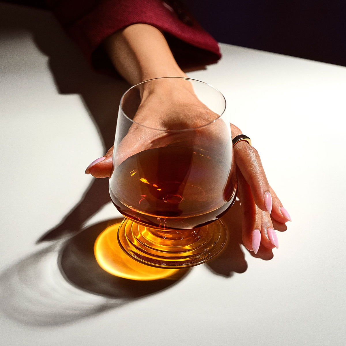 Overhead close-up of a hand holding the NUDE | Tom Dixon Splash Lowball glass filled with amber liquid, warm light casting long shadows across a white surface.