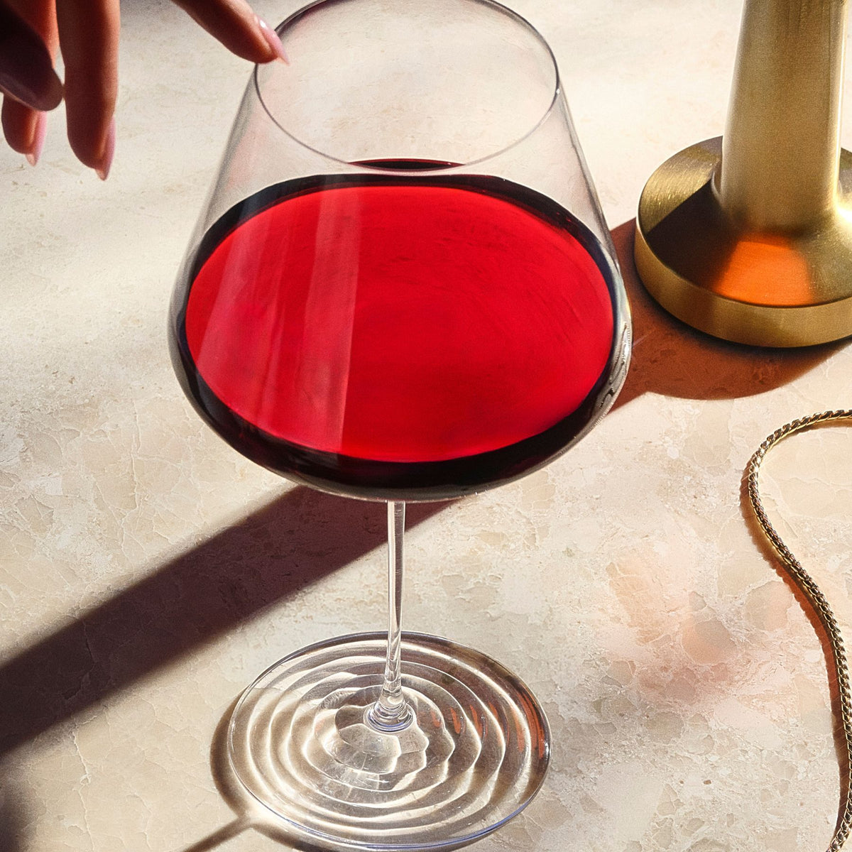 Overhead close-up of the NUDE Splash Red Wine glass filled with red wine, its ripple foot base visible, next to a gold candlestick and chain necklace on a warm marble surface, with a hand reaching into frame.