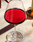 Overhead close-up of the NUDE Splash Red Wine glass filled with red wine, its ripple foot base visible, next to a gold candlestick and chain necklace on a warm marble surface, with a hand reaching into frame.