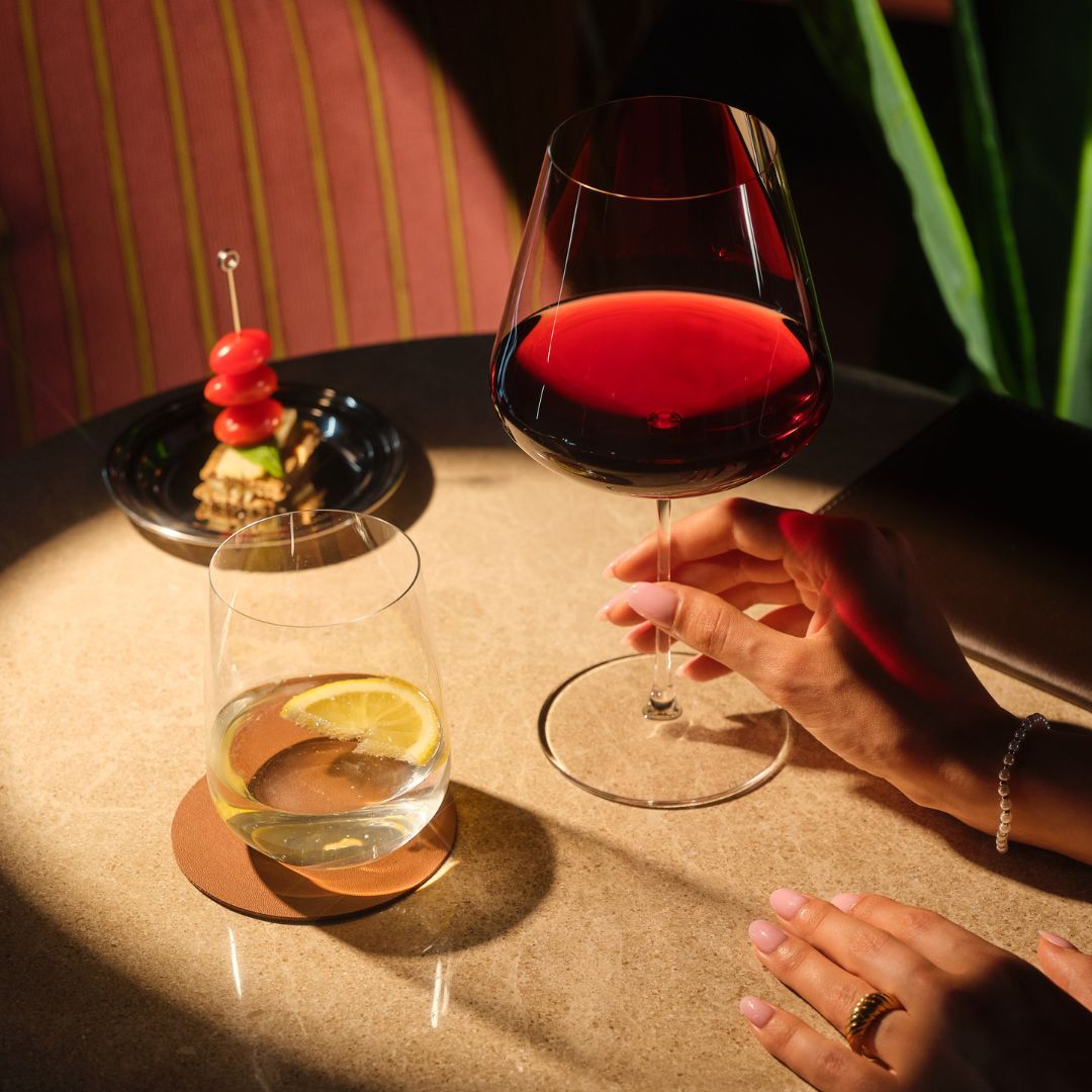 A hand holds a Sera Burgundy glass of red wine beside a Sera Tumbler of water and lemon, with a skewered appetizer on a small plate nearby.