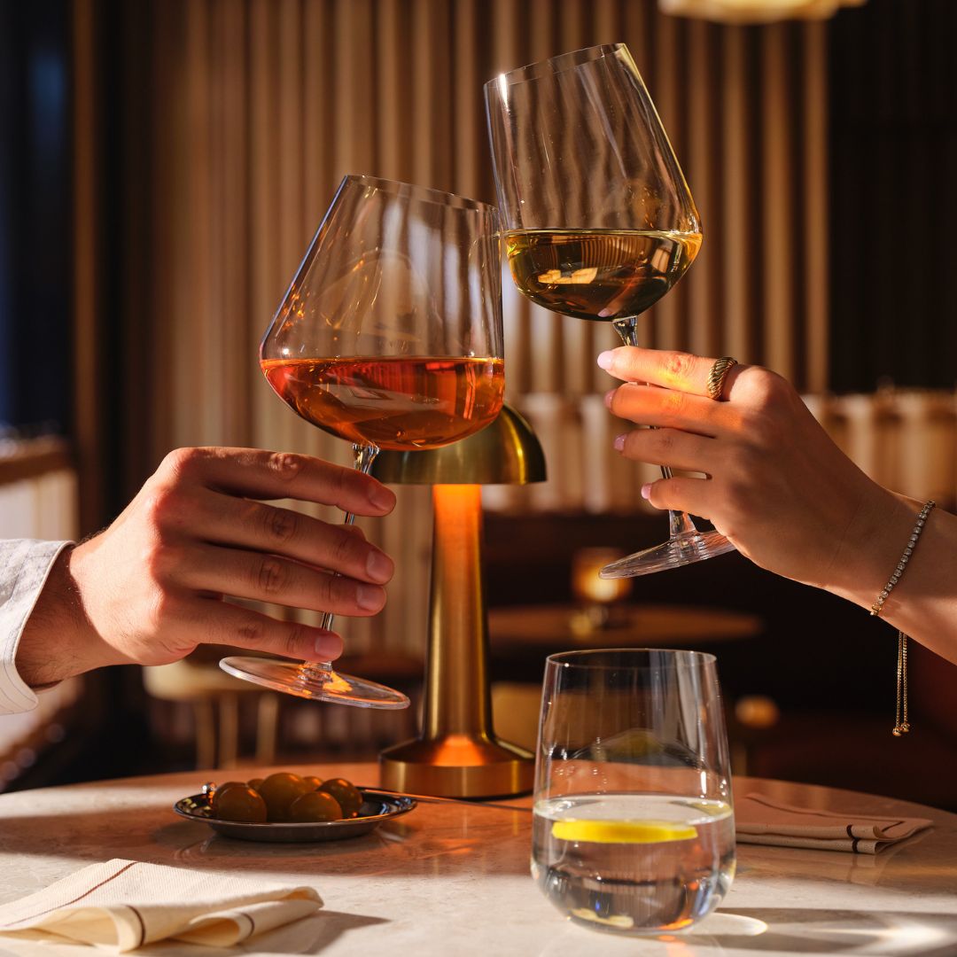 Two hands clink Sera Burgundy and Multipurpose wine glasses filled with orange and white wine, with a Sera Tumbler of water and lemon on the table below.
