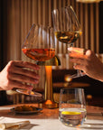 Two hands clink Sera Burgundy and Multipurpose wine glasses filled with orange and white wine, with a Sera Tumbler of water and lemon on the table below.