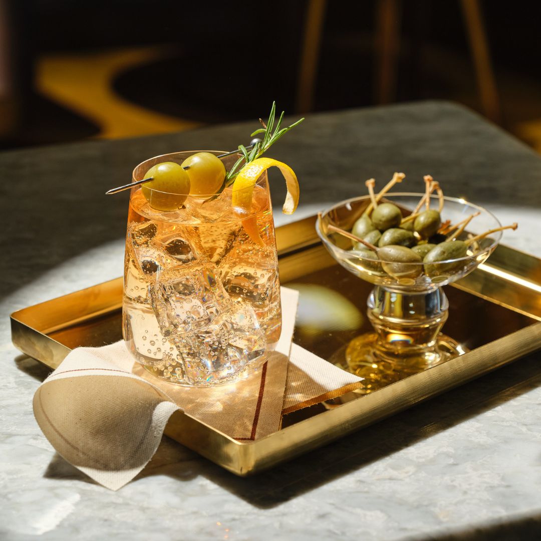 A styled cocktail in a Sera tumbler glass with ice, garnished with green olives, a lemon twist, and a rosemary sprig, served on a gold tray alongside a small glass Silhouette bowl of green olives on a stone tabletop.