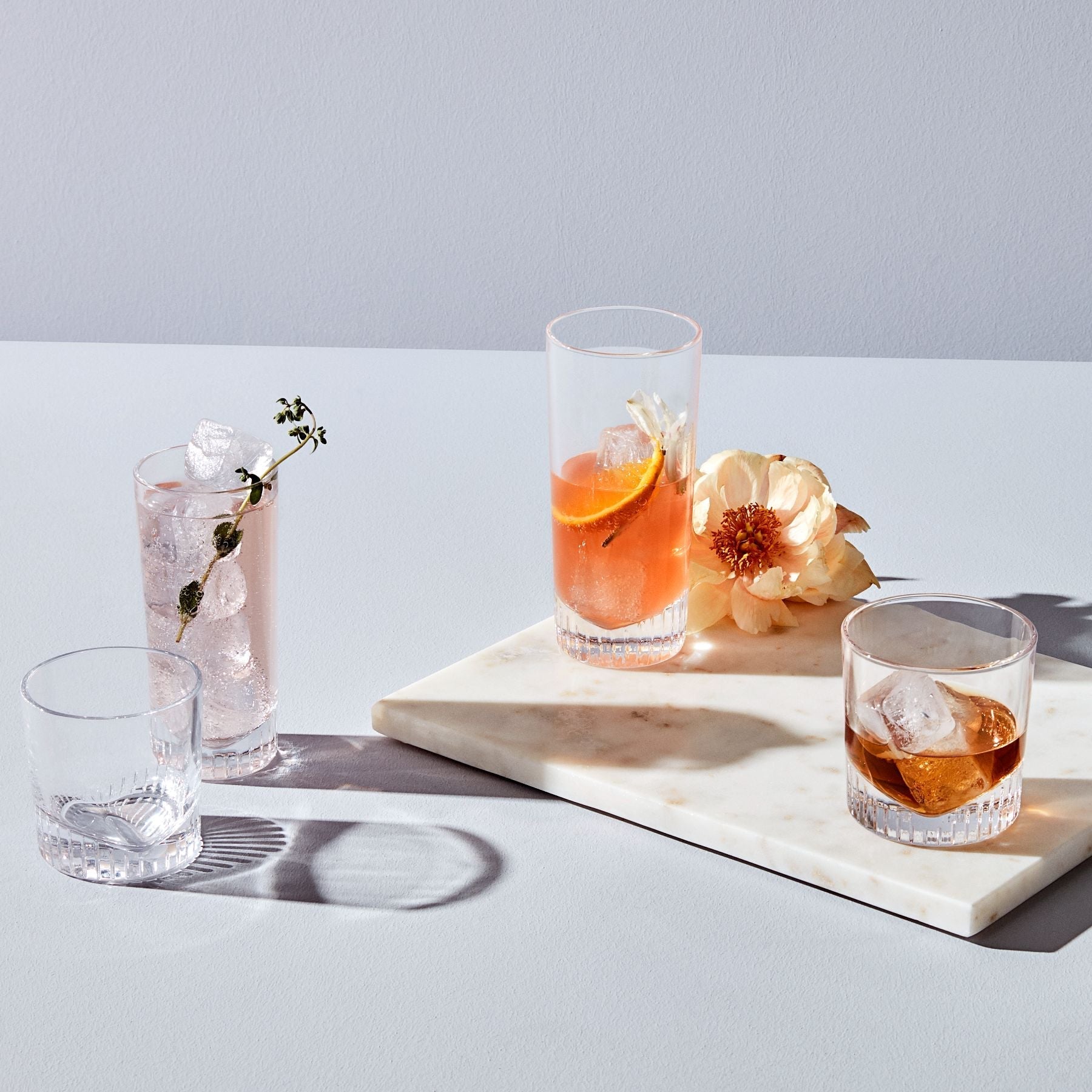 NUDE Caldera collection, lowball and highball glasses with v-shaped heavy bottoms, presented as a group filled with soft colored cocktails on a pink background