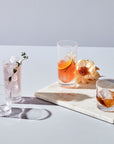 NUDE Caldera collection, lowball and highball glasses with v-shaped heavy bottoms, presented as a group filled with soft colored cocktails on a pink background