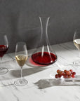 Cone shaped tall wine decanter in lead-free crystal, presented with red wine in it on a table with tablecloth surrounded by white, red and champagne glasses which are filled